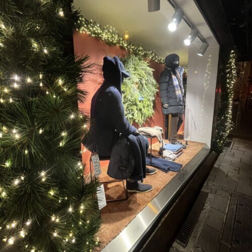 Schaufenster mit winterlicher Dekoration, Tannen, und Lichtern; zwei Schaufensterpuppen in Mänteln.