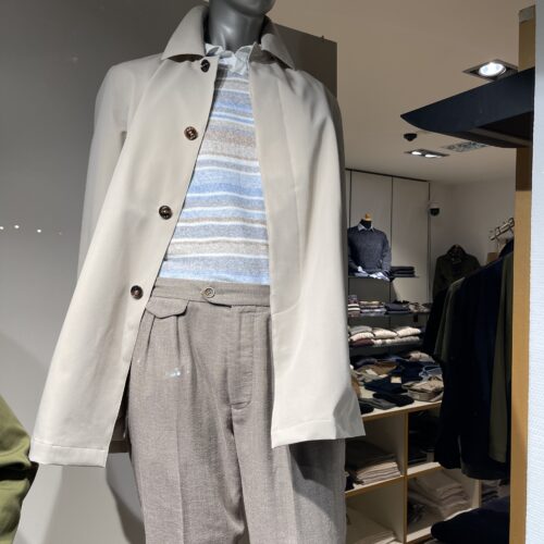 Eine Schaufensterpuppe mit beiger Jacke und gestreiftem Pullover in einer Ladenauslage.