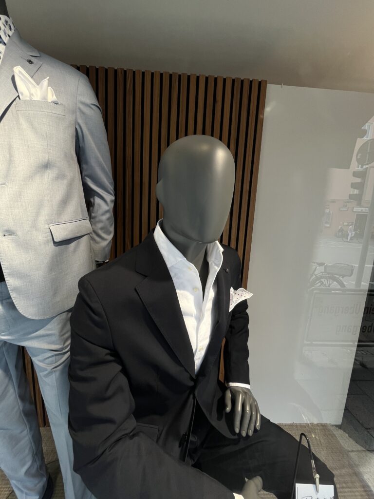 Eine Schaufensterpuppe im schwarzen Anzug und weißem Hemd mit Einstecktuch sitzt elegant