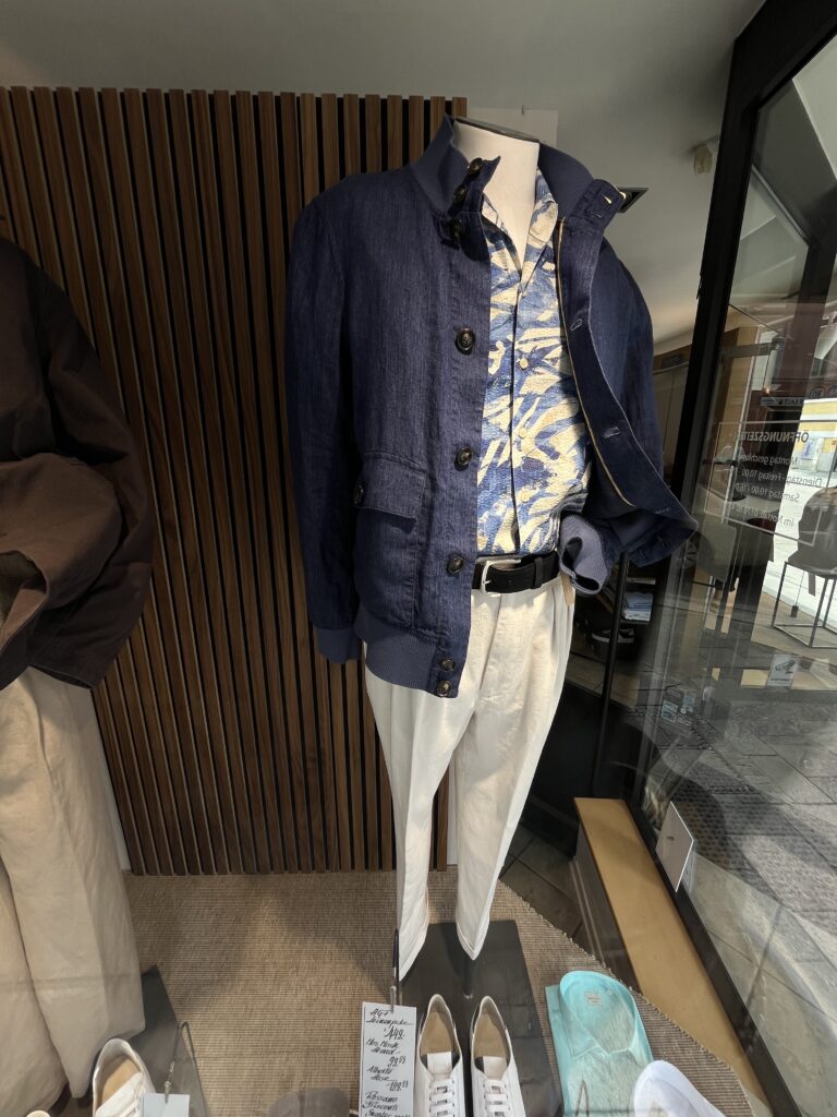 Eine männlich wirkende Schaufensterpuppe trägt ein blaues Hemd mit Muster, helle Hose und Jacke